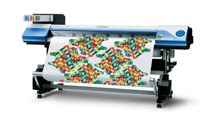 A large industrial print machine with a blue and silver body, shown printing a colourful geometric pattern onto a wide roll of white material. The machine is positioned on a clean white background with a soft shadow underneath.